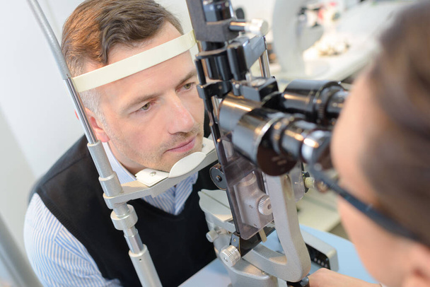 optometrist in exam room with man in chair - Fotó, kép