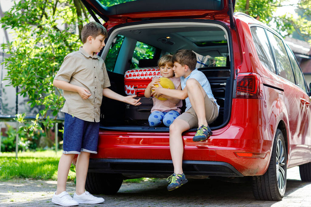 Three children, two boys and preschool girl sitting in car trunk before leaving for summer vacation with parents. Happy kids, siblings, brothers and sister with suitcases and toys going on journey - Valokuva, kuva