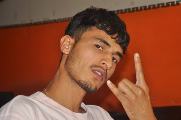 Close-up van een Zuid-Aziatische jongeman tonen rock hand gebaar met zijn hand, binnen zitten met het kijken naar de camera - Foto, afbeelding