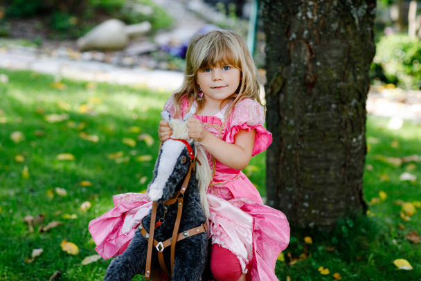 Little preschool girl hugging with rocking horse toy. Happy child in princess dress on sunny summer day in garden. Girl in love with her favourite old vintage toy animal. - Photo, Image