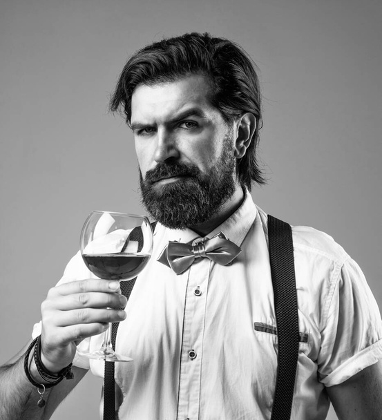 elegant male barman. handsome hipster drinking wine glass. sommelier tasting alcohol. bearded man in suspenders drink red wine. brutal guy bartender in bow tie - Zdjęcie, obraz