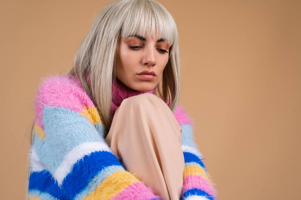 A blonde with long hair and bangs, with nude makeup and in a multi-colored bright fluffy sweater on a beige background, sad looks nowhere - Photo, Image