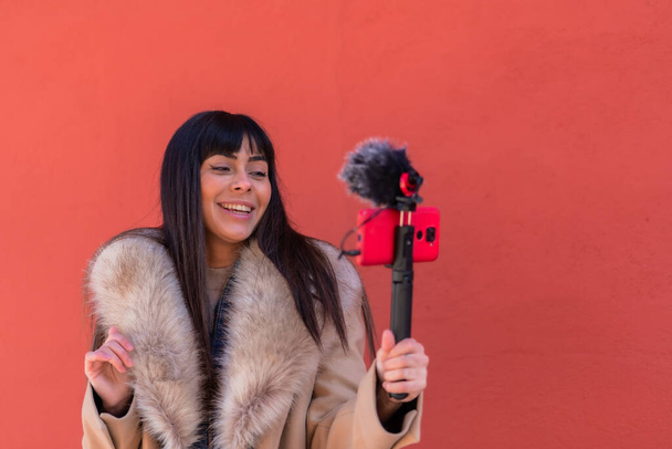Smiling brunette girl recording a video blog with her mobile phone and microphone, wearing a winter jacket for social networks. Caucasian pretty a red background - Photo, Image