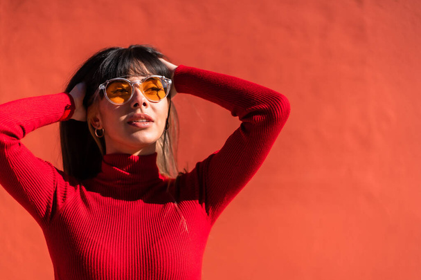 Posing brunette girl smiling in a dress and orange sunglasses in spring, enjoying a sunny morning. Caucasian pretty a red background, copy space and paste - Foto, Imagen