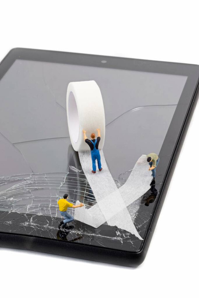 A team of miniature workmen repairing a broken mobile phone tablet touchscreen with a roll of sticky tape - Photo, Image