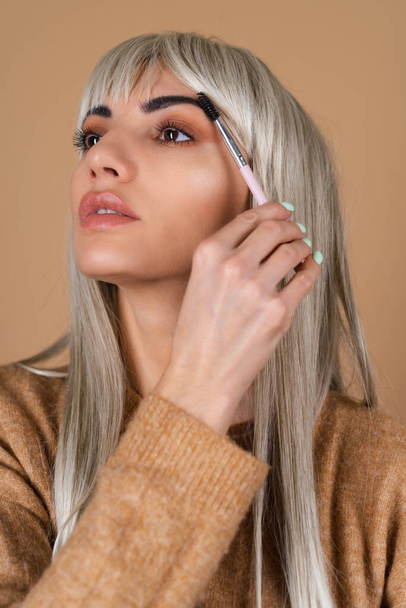 Блондинка с челкой и коричневым дневным макияжем держит кисть для бровей на бежевом фоне - Фото, изображение