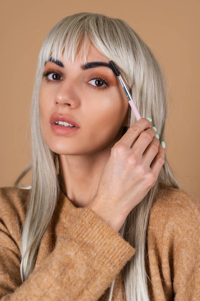 Blonde girl with bangs and brown daytime makeup holds an eyebrow brush on a beige background - Фото, зображення
