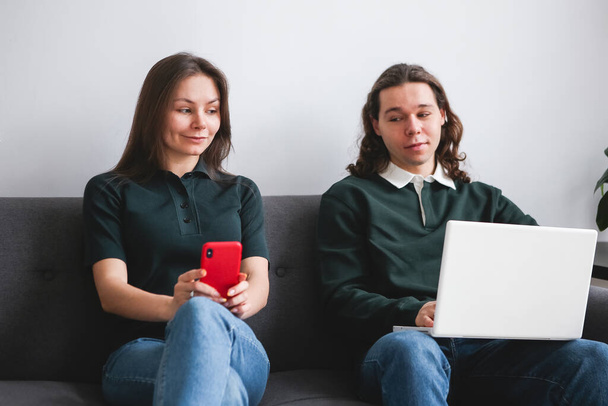 Couple sitting on the sofa with notebook and phone. Man and woman communicating on the electronic gadgets. Social isolation, distant learning students - Φωτογραφία, εικόνα