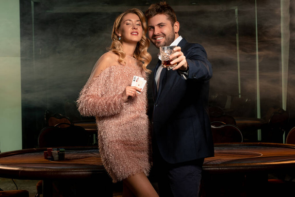 Žena a muž hráči pokeru stojící v kasinu drží pár esa a sklenice whisky - Fotografie, Obrázek