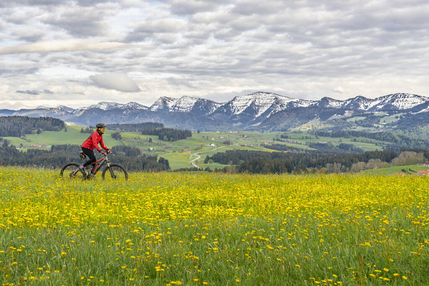 ciclismo de montaña de mujer mayor en una bicicleta de montaña eléctrica a principios de primavera, en el área de Allgaeu, una parte de los alpes bavarianos, Oberstaufen, Alemania, con la cadena montañosa Nagelfluh en el fondo - Foto, Imagen