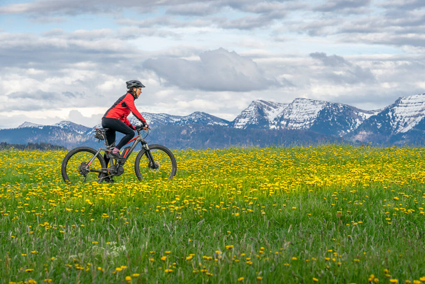 mulher sênior montanhismo em um e-mountain bike no início da primavera, na área de Allgaeu, uma parte dos alpes bávaros, Oberstaufen, Alemanha, com a cadeia de montanha Nagelfluh no fundo - Foto, Imagem