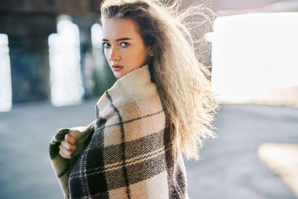 Beautiful young woman portrait wrapped in wool blanket looking to camera on windy sunny day on urban street background - Foto, Imagem