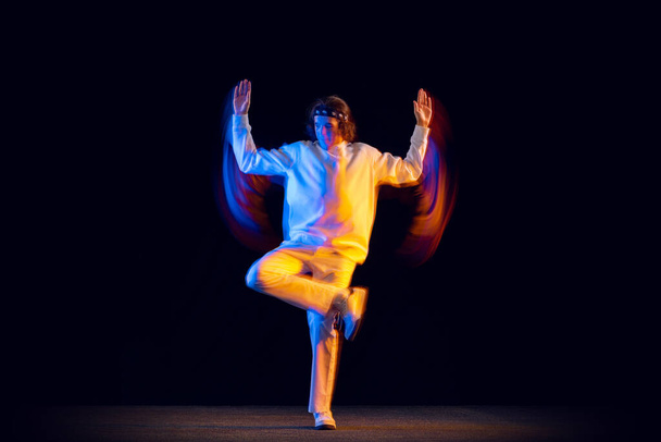 Homme élégant en costume blanc sportif dansant hip-hop isolé sur fond sombre dans la lumière au néon mixte. Culture jeunesse, hip-hop, mouvement, style et mode, action. - Photo, image