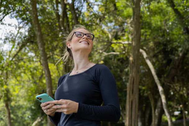 Nuori nainen käyttää älypuhelinta päivällä, taustalla vihreä puisto. Laadukas kuva. Matkapuhelin, teknologia, kaupunkikonsepti. Laadukas kuva - Valokuva, kuva