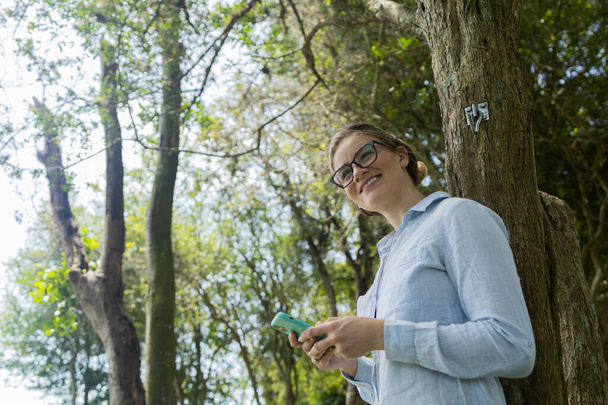 Mujer joven usando un teléfono inteligente durante el día con un parque verde en el fondo. Foto de alta calidad. Teléfono móvil, tecnología, concepto urbano. Foto de alta calidad - Foto, Imagen