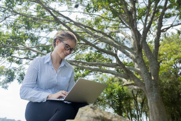 Νεαρή γυναίκα που χρησιμοποιεί φορητό υπολογιστή την ημέρα με ένα πράσινο πάρκο στο παρασκήνιο. Υψηλής ποιότητας φωτογραφία. Κινητό τηλέφωνο, τεχνολογία, αστική έννοια.  - Φωτογραφία, εικόνα