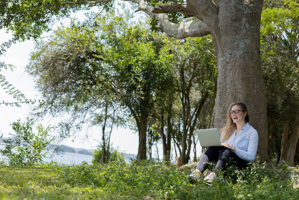 Молода жінка використовує ноутбук вдень з зеленим парком на задньому плані. Якісна фотографія. Мобільний телефон, технології, міська концепція
.  - Фото, зображення