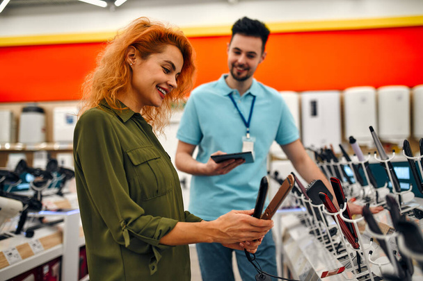 Ένα όμορφο κορίτσι με κόκκινα μαλλιά μιλάει σε έναν άντρα πωλητή ενώ παίρνει βοήθεια αγοράζοντας ένα νέο μηχάνημα για τα μαλλιά σε ένα κατάστημα σιδηρικών και ηλεκτρονικών ειδών. - Φωτογραφία, εικόνα
