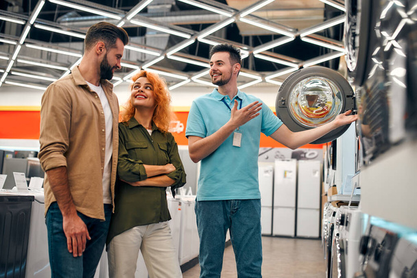 A beautiful red-haired girl and a bearded man buy a washing machine in a home appliances and electronics store. A male salesman helps a couple choose new appliances for their home. - Fotografie, Obrázek