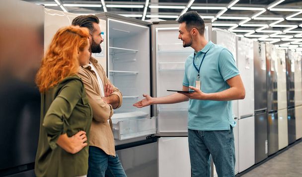 Beautiful red-haired woman and bearded man buying a refrigerator in a home appliances and electronics store. Male sales assistant helping a couple choose new appliances for the home. - Photo, Image