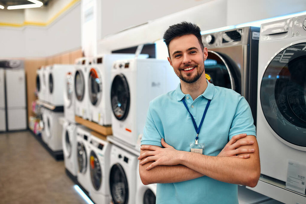 Un joven asistente de ventas barbudo en uniforme de pie con los brazos cruzados en una tienda de electrodomésticos y electrónica contra el telón de fondo de lavadoras. - Foto, Imagen