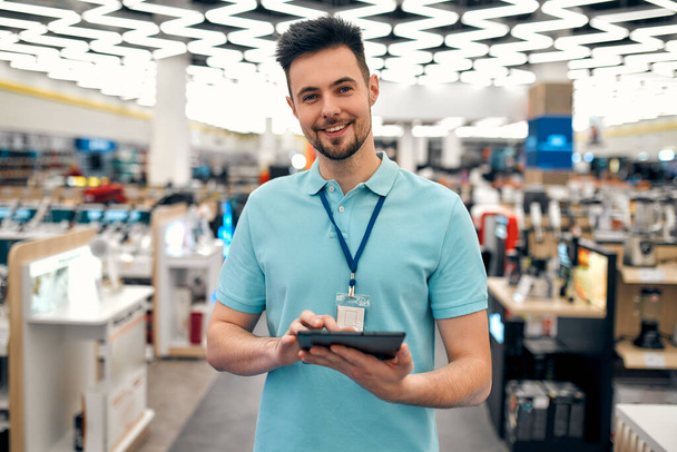 Un joven asistente de ventas exitoso en uniforme con una tableta en sus manos contra el fondo del interior de la sala en una tienda de electrodomésticos y electrónica. - Foto, imagen