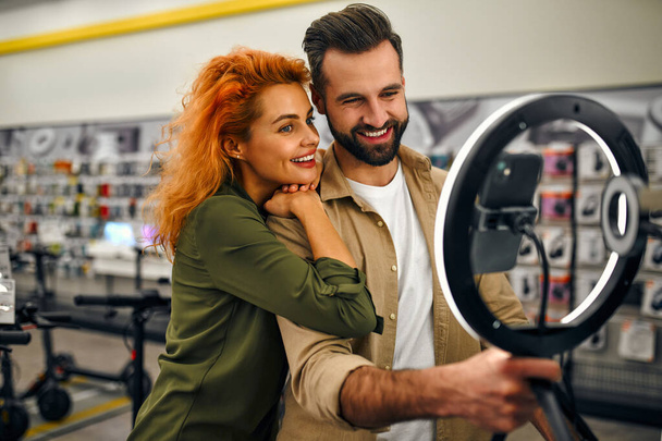 A young red-haired woman with a bearded man take a selfie with a ring led lamp in a gadget and electronics store. - Zdjęcie, obraz