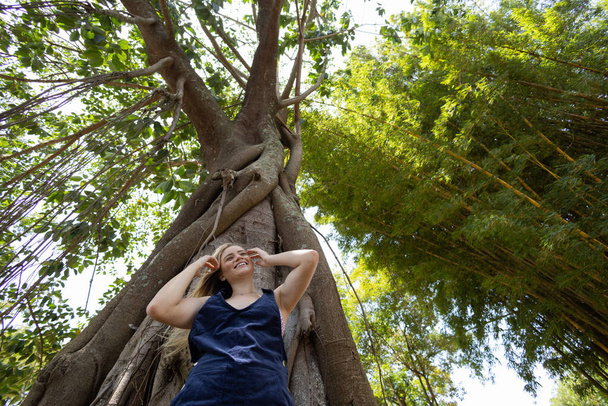 Retrato de una joven sonriente en un parque en un hermoso día soleado. Mujer con los brazos abiertos frente a un árbol grande. Fondo verde y naturaleza. Foto de alta calidad - Foto, imagen