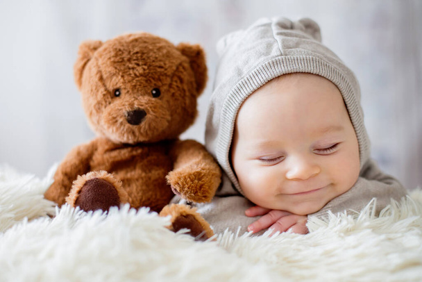 Newbor baby boy in bear overall, happily smiling, sleeping in bed with teddy bear stuffed toys, pure white, isolated, selective focus.  - Photo, image