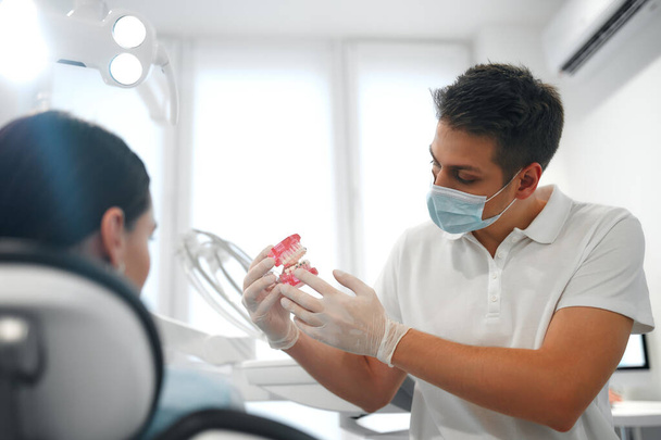 Jonge tandarts in wit uniform en beschermende medische masker tonen kaak model met beugel aan vrouwelijke patiënt in tandheelkunde kantoor met moderne apparatuur. - Foto, afbeelding