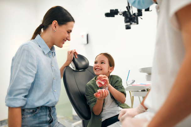 Child little cute girl with her mom sitting on a dental chair holding a model of jaws. Pediatric dentist explaining to the patient about dental care. - Fotografie, Obrázek
