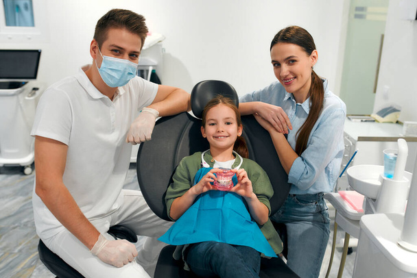 Przyjemna dziewczynka leży na fotelu dentystycznym i trzyma model szczęk, obok niej jest jej matka i dentysta dziecięcy. Stomatologia i opieka dentystyczna. - Zdjęcie, obraz