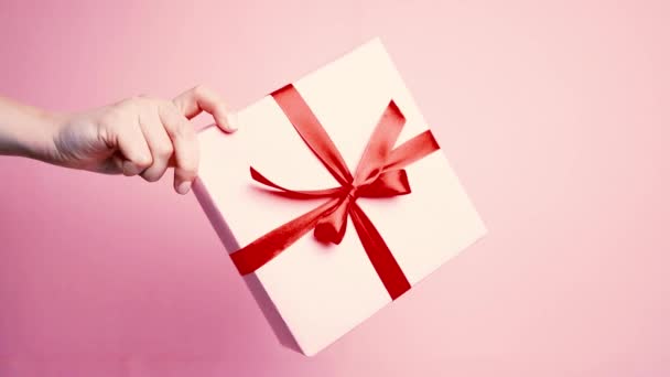 Kaukasische Hand zeigt Überraschung Geschenkbox mit roter Schleife Schleife isoliert auf rosa Pastell Hintergrund - Filmmaterial, Video