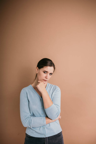 een twijfelende brunette vrouw in een blauwe sweater staat en kijkt naar de camera op een beige achtergrond - Foto, afbeelding