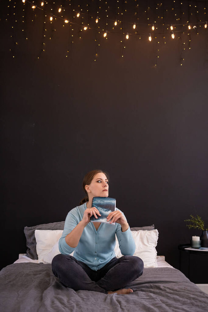 Eine traurige brünette Frau sitzt auf einem Bett in einem Zimmer mit einem Buch. Angenehme Freizeit - Foto, Bild