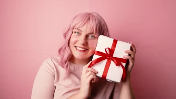 Juhlitaan. Onnellinen positiivinen nuori valkoihoinen nainen, joka näyttää lahjapakkausta, kun katsot kameraa, joka on eristetty vaaleanpunaisella taustalla - Materiaali, video