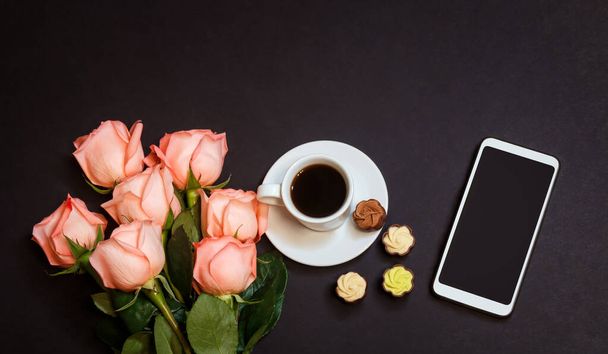 Pembe güller, kahve fincanı, akıllı telefon ve koyu arkaplanda çikolata şekeri. Üst görünüm, alanı kopyala. - Fotoğraf, Görsel