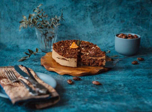 Homemade cake with chocolate and cream on round wooden board next to a plate with a fork and knife and a mug filled with nuts on dark background. Home baking - Φωτογραφία, εικόνα