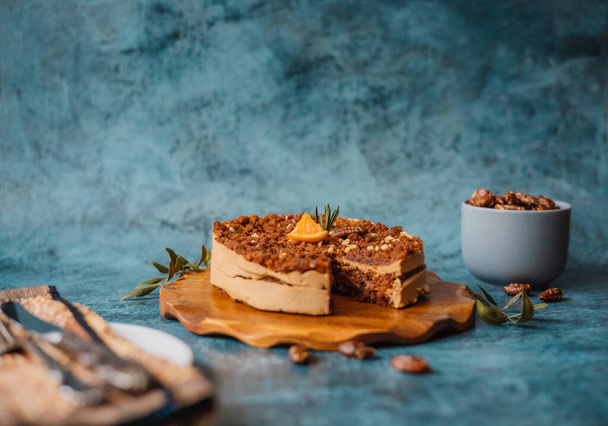 Homemade cake with chocolate and cream on round wooden board next to a plate with a fork and knife and a mug filled with nuts on dark background. Home baking - 写真・画像