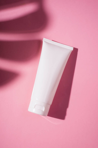 Plastic white tube for cream or lotion. Skin care or sunscreen cosmetic in top view on pink background with palm leaves shadow. Beauty concept for face care - Zdjęcie, obraz
