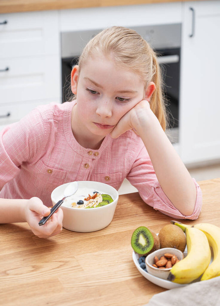 Ein trauriges Mädchen in rosa Hemd sitzt am Küchentisch und isst Joghurt mit Müsli und frischem Obst. Das Konzept der gesunden Ernährung. - Foto, Bild