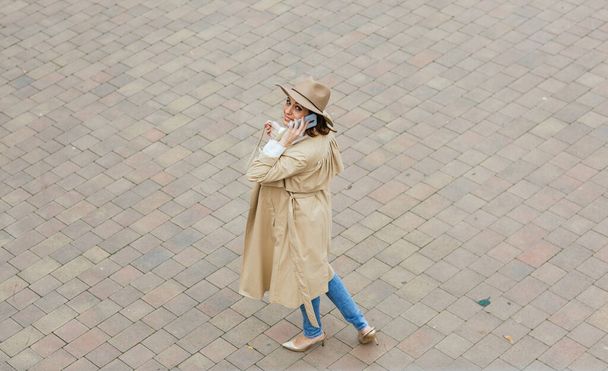 Vrouw die naar de camera kijkt en aan de telefoon praat terwijl ze op straat loopt. - Foto, afbeelding