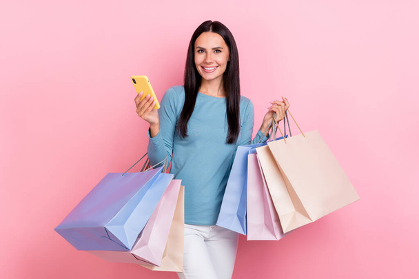 Photo of pretty adorable lady dressed blue shirt smiling holding shoppers typing modern device isolated pink color background - Photo, Image