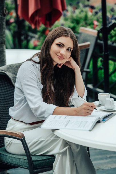 Smiling young dark-haired businesswoman makes notes in a notebook in a cafe on a summer day. Business, e-learning, freelance concept. laptop on a table - Foto, imagen