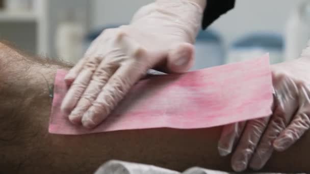 Procedimiento de depilación: el maestro frota la tira en la pierna de su cliente y rasga los pelos en la pierna - Imágenes, Vídeo