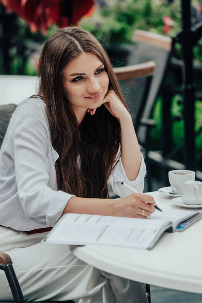 Die lächelnde junge dunkelhaarige Geschäftsfrau macht sich an einem Sommertag in einem Café Notizen in einem Notizbuch. Business, E-Learning, freiberufliches Konzept. Laptop auf einem Tisch - Foto, Bild