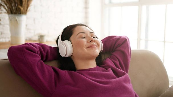 Young asian woman relaxing on comfortable sofa with eyes closed wearing headphones. Pretty mixed race lady enjoys listening chill music audio sound meditating feeling no stress at home. - Photo, image