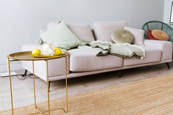 Υπάρχει ένας καναπές με μαξιλάρια μπροστά στον γκρίζο τοίχο, δίπλα στον καναπέ υπάρχει ένα διακοσμητικό τραπέζι σε χρυσό χρώμα. Υπάρχει ένας δίσκος με λεμόνια και ένα κηροπήγιο στο τραπέζι. - Φωτογραφία, εικόνα