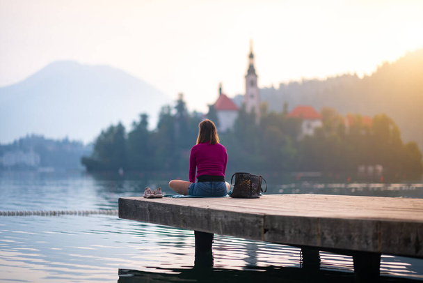 Lone girl sitting on a wooden jetty. Tranquil scene with meditating woman with the beautiful landscape in the background. - Fotografie, Obrázek