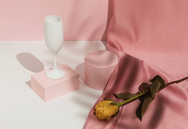 Ρομαντικό παστέλ ροζ σύνθεση Ημέρα του Αγίου Βαλεντίνου με ποτήρι κρασιού, σατέν κουρτίνα και κίτρινο τριαντάφυλλο λουλούδι. Κατάλληλο για Επίδειξη Προϊόντων και Business Concept. Σύγχρονη αισθητική. - Φωτογραφία, εικόνα